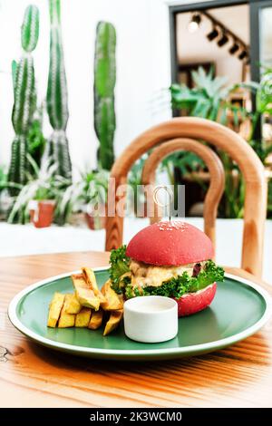 Veganer Burger, in einem roten Brötchen, Gericht mit Chips, auf einem grünen Teller, auf einem Holztisch, gesundes vegetarisches Essen. Stockfoto