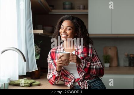Fröhliche tausendjährige afroamerikanische Frau mit Tasse Genießen Sie ein Getränk, Komfort und Ruhe, mit Blick aus dem Fenster Stockfoto