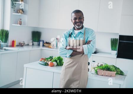 Foto von positiv gealterten Mann, der die Arme kreuzt, bereit, schmackhaftes Abendessen mit frischen Bio-Produkten im modernen Küchenhaus zu machen Stockfoto