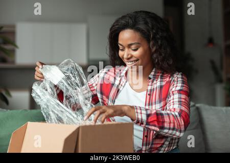 Eine fröhliche, tausendjährige afroamerikanische Frau öffnet einen Karton und packt das Paket im Wohnzimmer aus Stockfoto