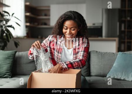 Die frohe tausendjährige afroamerikanische Frau öffnet einen Einkaufskarton mit Einkäufen und Auspacken des Pakets Stockfoto