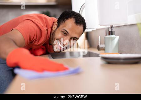 Lächelnder, tausendjähriger schwarzer Mann in Gummihandschuhen wischt Staub vom Holztisch im Kücheninnenraum, freien Platz Stockfoto