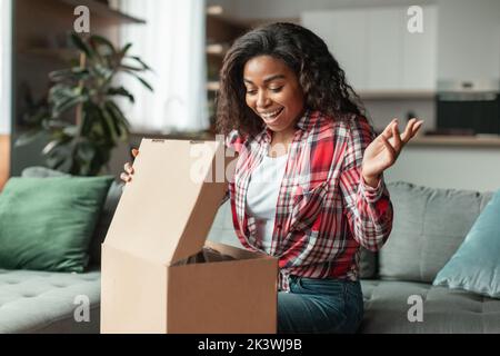 Schockiert glücklich tausendjährige afroamerikanische Frau öffnet Karton schaut auf Einkäufe, genießen Verkauf Stockfoto