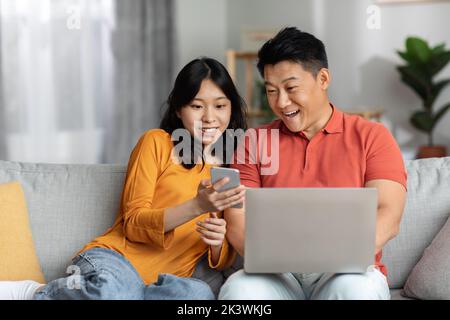 Positive japanische Mann und Frau sitzen auf der Couch, mit Gadgets Stockfoto