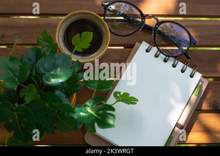 Kaffeetasse, Notizbuch und Pflanzen auf Holztisch. Kaffee Espresso an sonnigen Tagen im Café. Flacher Schreibtisch mit Kaffee, grüner Pflanzenpalme, Brille. Top Vie Stockfoto