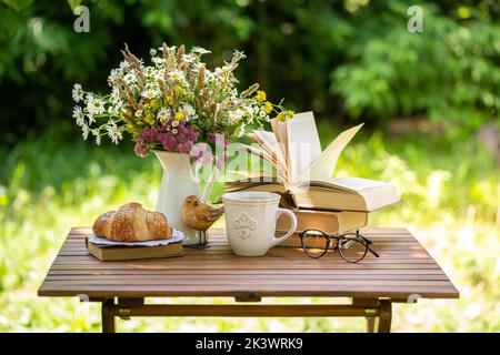 Strauß Wiesenblumen, Croissant, Tasse Tee oder Kaffee, Bücher auf dem Tisch im idyllischen Sommergarten. Erholung im Garten, Bücher lesen, Frühstück, vacatio Stockfoto