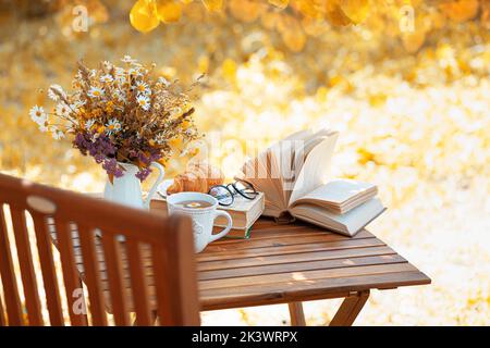 Blumenstrauß, Croissant, Tasse Tee oder Kaffee, Bücher auf dem Tisch im Herbstgarten. Erholung im Garten, Bücher lesen, Frühstück, Urlaub in der Natur Co Stockfoto