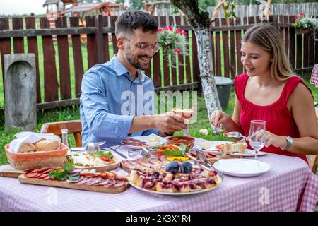 Ein glückliches junges romantisches Paar, das Mittag- oder Abendessen in einem wunderschönen Open-Air-Landrestaurant mit traditionellem Essen hat. Stockfoto