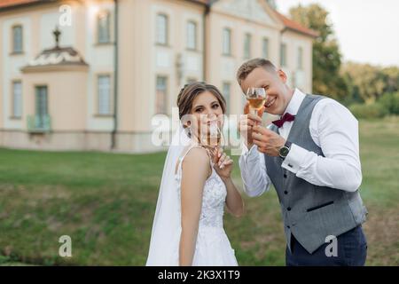 Portrait eines erstaunlichen Ehepaares, das im Sommer in der Nähe eines alten Gebäudes im Park steht und mit Weingläsern die Hände hebt. Stockfoto