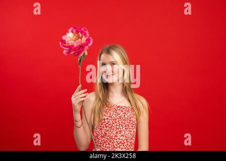 Portrait von fröhlichen erstaunlich Teenager-Mädchen mit schönen Haaren halten Heben Hand mit Blume rosa Pfingstrose auf rotem Hintergrund. Stockfoto