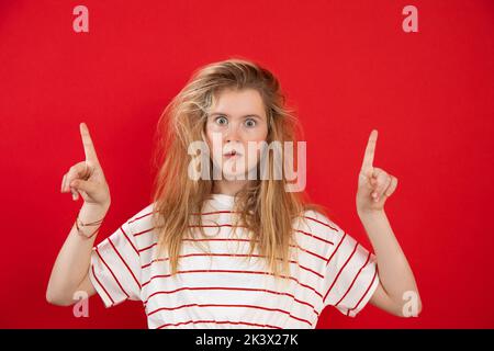 Porträt eines schockierten attraktiven Teenagers, das weißes T-Shirt mit roten Streifen trägt und die Hände mit den Zeigefinger nach oben hebt. Stockfoto