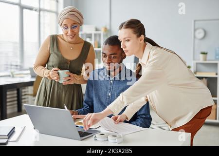 Eine vielfältige Gruppe von drei Geschäftsleuten, die gemeinsam mit einem Laptop über das Projekt im Minimalbüro sprechen Stockfoto