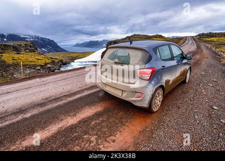 Auto voller rotem Schlamm auf einer schlammigen Schotterstraße in Island bei bewölktem Wetter. Typische isländische Landschaft im Winter im Hintergrund. Stockfoto