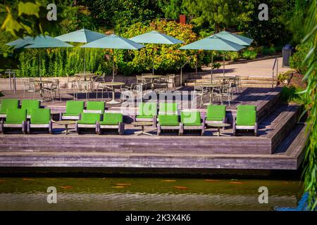 Große Holzterrasse am Wasser mit Tischen und Sonnenschirmen und grün gepolsterten rustikalen Schaukelstühlen - üppiges Laub im Hintergrund und große orange Koi schwimmen Stockfoto