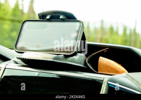 Mobiltelefon in einer gewichteten Clamshell-Klammer und Sonnenbrillen-Hülle auf dem Armaturenbrett des Fahrzeugs mit verschwommenen Bäumen in der Windschutzscheibe - Weiches Scharfstellen Stockfoto