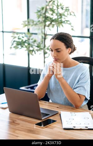 Vertikales Foto einer nachdenklichen, erfolgreichen brasilianerin oder Latinerin, Büroangestellter, Kreativmanagerin, sitzt am Arbeitsplatz im modernen Büro, schaut auf einen Laptop, denkt an ein Projekt, plant Stockfoto
