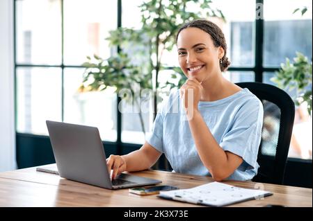 Selbstbewusst erfolgreiche elegante, hübsche brasilianische oder hispanische junge Frau, geschäftsführerin, Produkt- oder Finanzmanagerin, in blauem Hemd, sitzt am Schreibtisch im modernen Büro, blickt auf die Kamera und lächelt freundlich Stockfoto