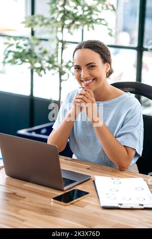 Vertikales Foto einer fröhlichen, eleganten, hübschen brasilianerin oder Latino-Frau, Büroangestellter, Produkt- oder Finanzmanagerin, sitzt am Schreibtisch in einem modernen Büro, schaut freundlich lächelnd auf die Kamera Stockfoto