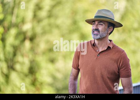Mann mittleren Alters, der einen Hut trägt und wegschaut, während er im Freien vor einem natürlichen Hintergrund steht. Stockfoto
