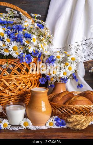 Rustikales Stillleben. Milch und Brot mit Blumen. Kamillen und Kornblumen in einem Korb Stockfoto