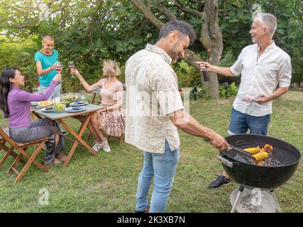 Freunde, die Bier trinken und mit dem Grill kochen, lächeln glücklich und lachen gemeinsam auf der Terrasse im Freien. Stockfoto