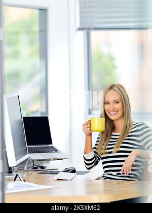 Ich liebe meinen Job. Eine schöne junge Frau, die an ihrem Schreibtisch arbeitet, während sie einen Drink genießt. Stockfoto