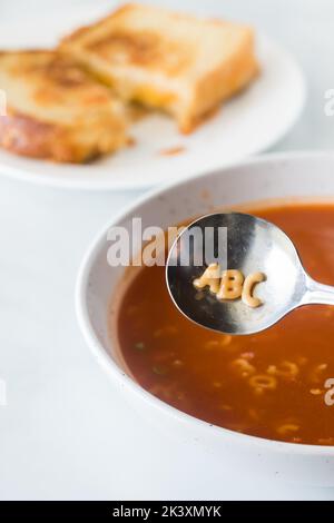 Alphabetsuppe mit den Buchstaben A, B, C im Löffel und gegrilltem Käse hinten Stockfoto