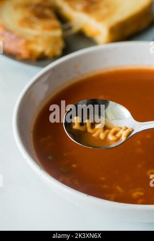 Eine Schüssel mit Tomaten-Alphabet-Suppe mit dem Wort Mittagessen im Löffel. Stockfoto