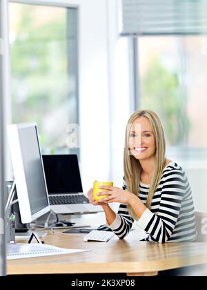 Eine schöne junge Frau, die an ihrem Schreibtisch arbeitet und dabei einen Drink genießt. Stockfoto