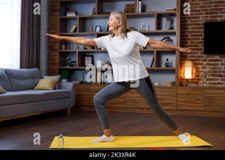 Grauhaarige, glückliche, reife Frau, die online mit dem Laptop Gymnastik und Yoga zu Hause macht. Aktiver und gesunder Lebensstil für Rentner. Fitnesskurse Stockfoto