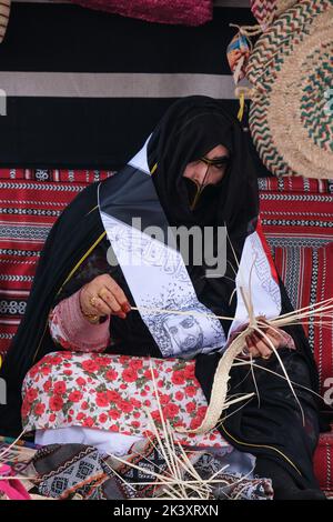 Maskierte arabische Frau aus Abu Dhabi, die eine Abaya trägt und Bands aus Dattelpalmfaser für die Herstellung traditioneller Strohkörbe herstellt. Stockfoto