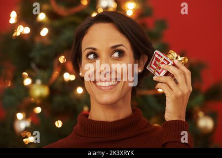 Ich hoffe, es ist das, was ich wollte. Eine attraktive junge Frau, die ein Geschenk hält, während sie an einem Weihnachtsbaum steht. Stockfoto