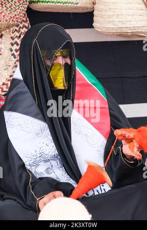 Maskierte arabische Frau aus Abu Dhabi trägt eine Abaya bei einem Folklife Festival, das traditionelle Handwerkskunst demonstriert und Wolle auf eine Spindel setzt. Stockfoto