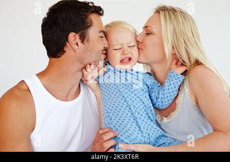 Ihn mit Küssen ersticken. Zwei liebevolle Eltern küssen ihren Sohn auf seine Wangen, während er aufgeregt aussieht und versucht, sie wegzustoßen. Stockfoto