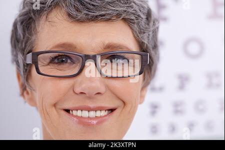 Ich kann jetzt klar sehen. Nahaufnahme einer reifen Frau mit Brille, die vor einem Augentestplakat steht. Stockfoto