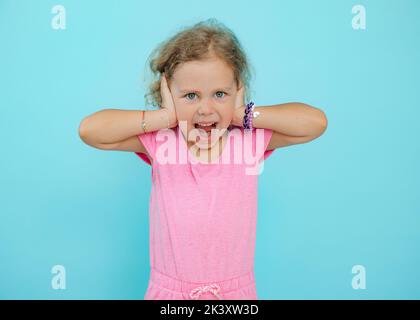 Porträt eines erstaunlichen blauäugigen kleinen Mädchens, das die Kamera anschaut, die Ohren mit Händen bedeckt und auf blauem Hintergrund schreit. Stockfoto