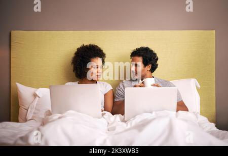 Arbeitet, während er sich unterhält. Ein junges Paar im Bett mit ihren Laptops. Stockfoto