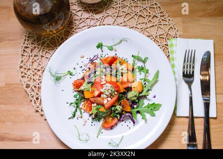 Warmer Salat mit Kürbis, Tomaten, Rucola, Pesto-Sauce mit Pinienkernen und schwarzen Sesam auf weißem Teller Draufsicht Stockfoto