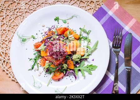 Warmer Salat mit Kürbis, Tomaten, Rucola, Pesto-Sauce mit Pinienkernen und schwarzen Sesam auf weißem Teller Draufsicht Stockfoto