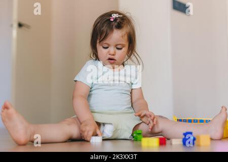 Kleines Mädchen, das auf dem Boden sitzt und im Wohnzimmer mit vielen bunten Holzblöcken spielt. Genießen Sie die Freizeit in Stockfoto