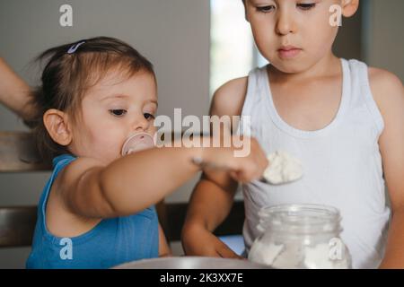 Zwei kleine Kinder, die Zutaten in die Schüssel geben und den Teig zu Hause am Tisch zubereiten. Menschen leben. Lifestyle-Konzept für die ganze Familie. Lebensmittelkonzept. Stockfoto