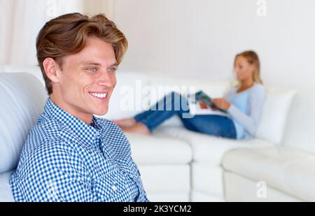 Einen faulen Sonntag zu Hause verbringen. Ein junger Mann, der sich zu Hause entspannt, während seine Freundin im Hintergrund ein Magazin liest. Stockfoto