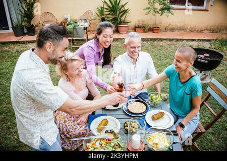 Erwachsene Freunde feiern einen Geburtstag und toasten Weingläser und Biere, die zusammen am Esstisch in einem Hinterhof des Hauses Spaß haben. Lifestyle-Konzept. Stockfoto