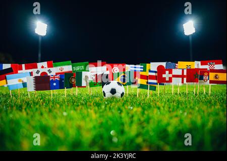 Konzeptfoto für internationales Fußballturnier. Ball auf grünem Gras und Nationalflaggen. Stadion in der Nacht. Schwarzer Hintergrund, Arbeitsbereich bearbeiten Stockfoto
