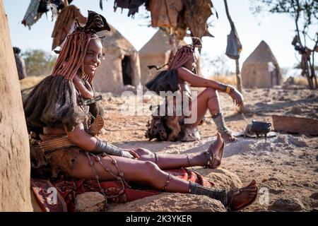 Himba-Frauen sitzen vor ihren Hütten in einem traditionellen Himba-Dorf in Namibia, Afrika. Stockfoto