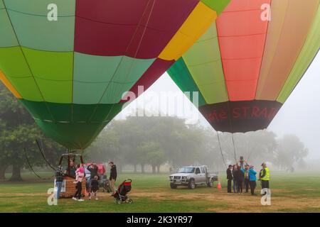 Zwei bunte Heißluftballons auf dem Boden an einem nebligen Tag, an dem Menschen um die Körbe stehen. „Balloons over Waikato“-Festival, Hamilton, Neuseeland Stockfoto