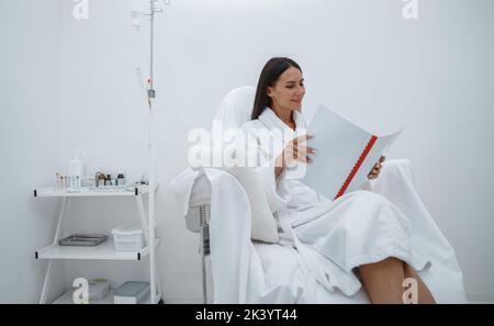 Schöne Frau im weißen Bademantel Lesemagazin, während sie im luxuriösen Wellness Spa-Center sitzt Stockfoto