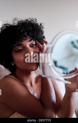 Selbstbewusste junge Transgender-Frau mit lockigen Haaren lächelt, während sie sich in einen kleinen Spiegel schaut. Stockfoto
