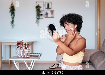 Transgender-Frau, die zu Hause Kompaktpuder auf ihr Gesicht aufgibt, während sie sich schminkt. Stockfoto
