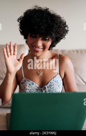 Transgender-Frau lächelt und winkt, während sie einen Videoanruf mit einem Laptop zu Hause auf einem Sofa sitzt. Stockfoto